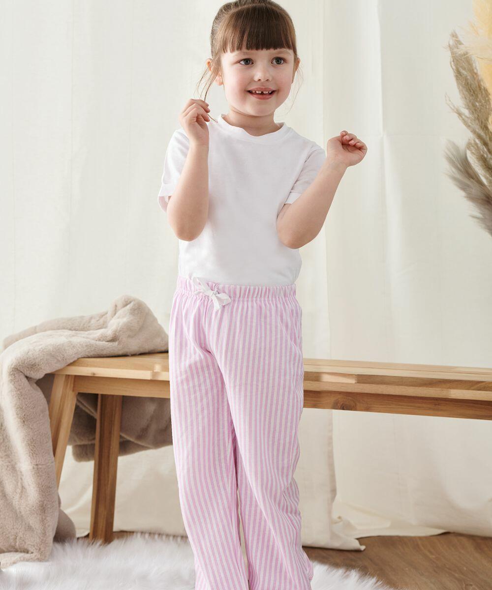 A child wearing a white shirt and pink striped pants stands indoors near a wooden bench, smiling and holding a small Towel City TC059 object in their hand.