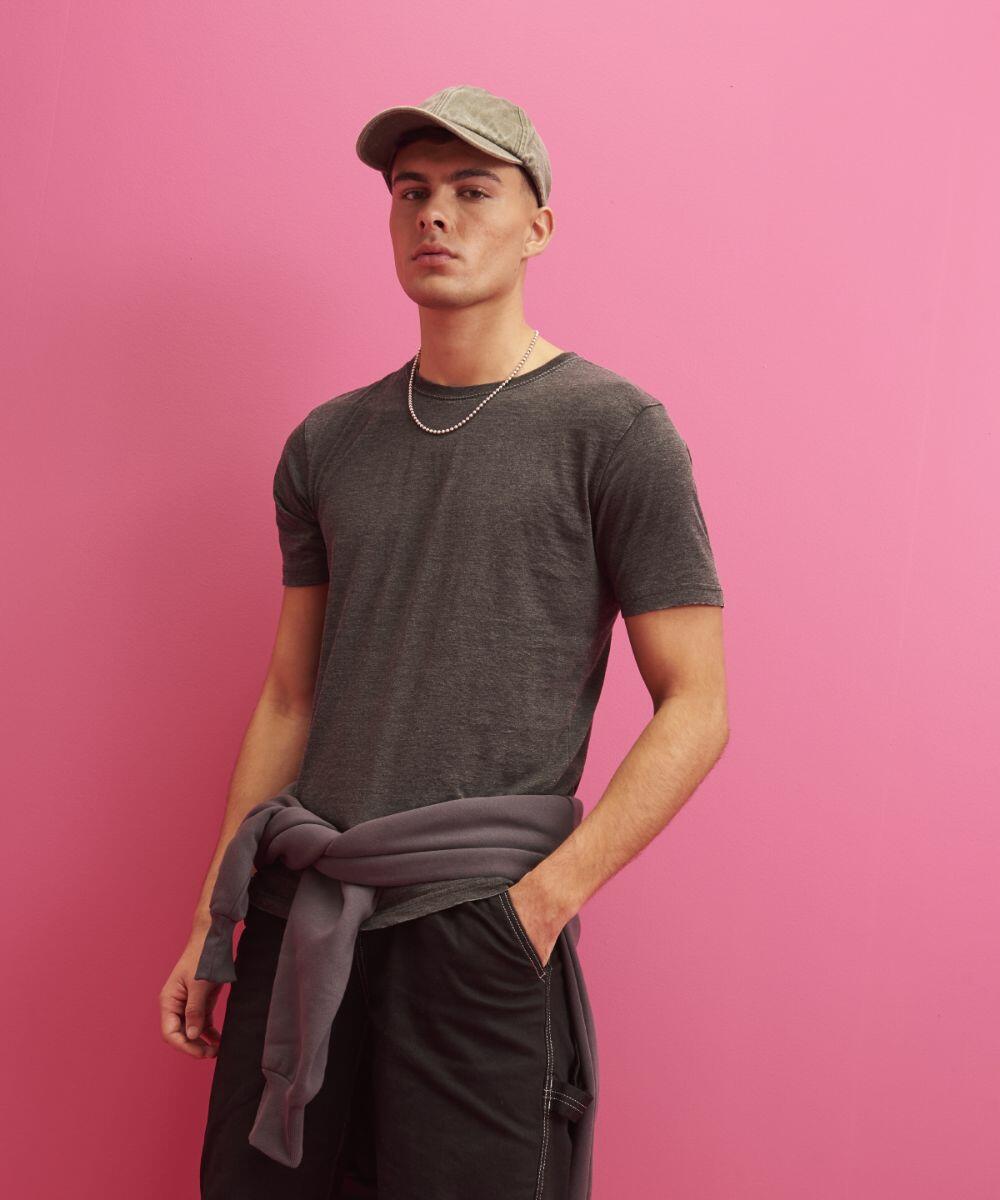 A man wearing an AWDis Just T's JT099 grey t-shirt, grey cap, and dark pants with a grey sweatshirt tied around his waist stands against a pink background.