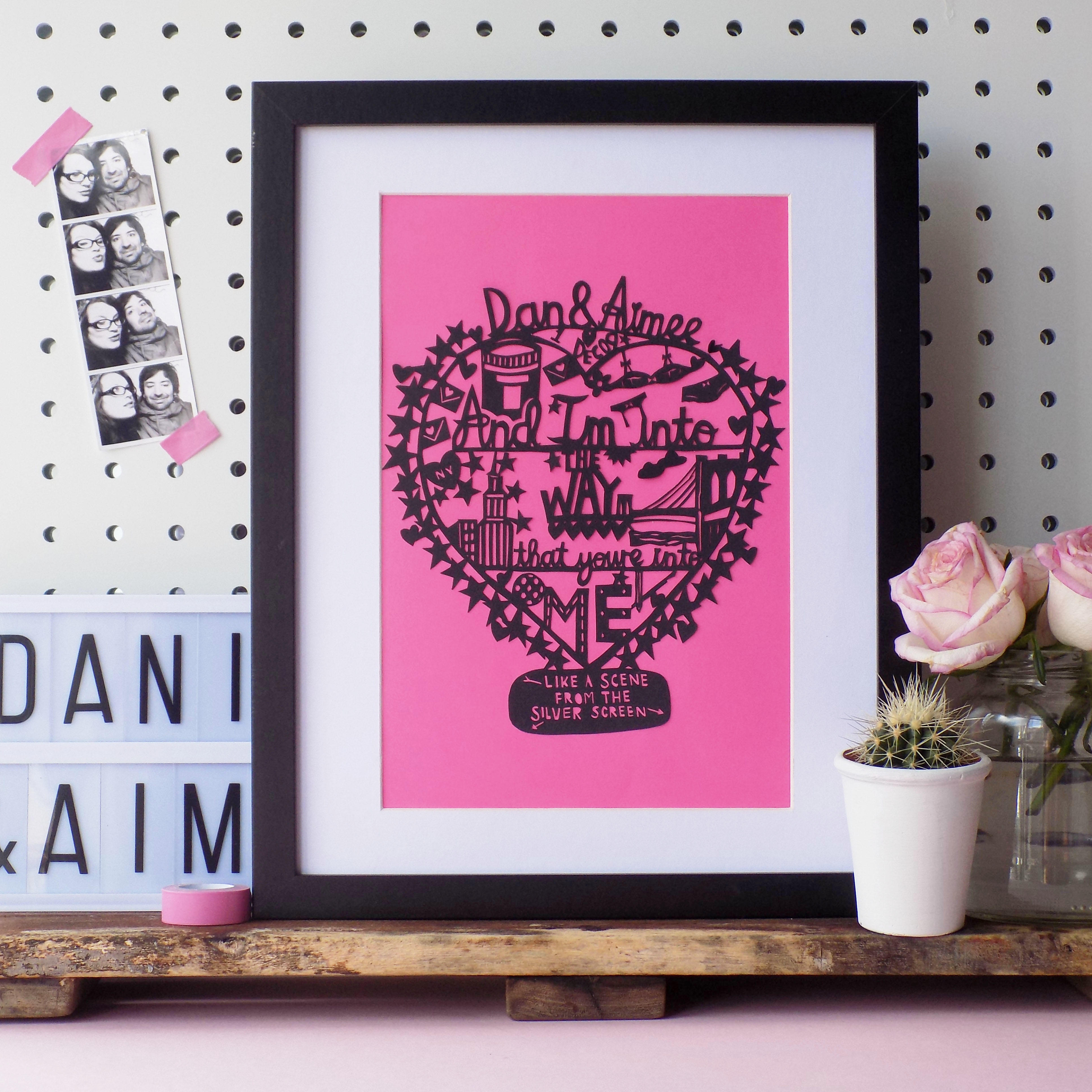 A hand drawn hand cut paper cut love heart featuring a song lyric, a special date, the couples names, and five small illustrations of their favourite things with a starry outline.