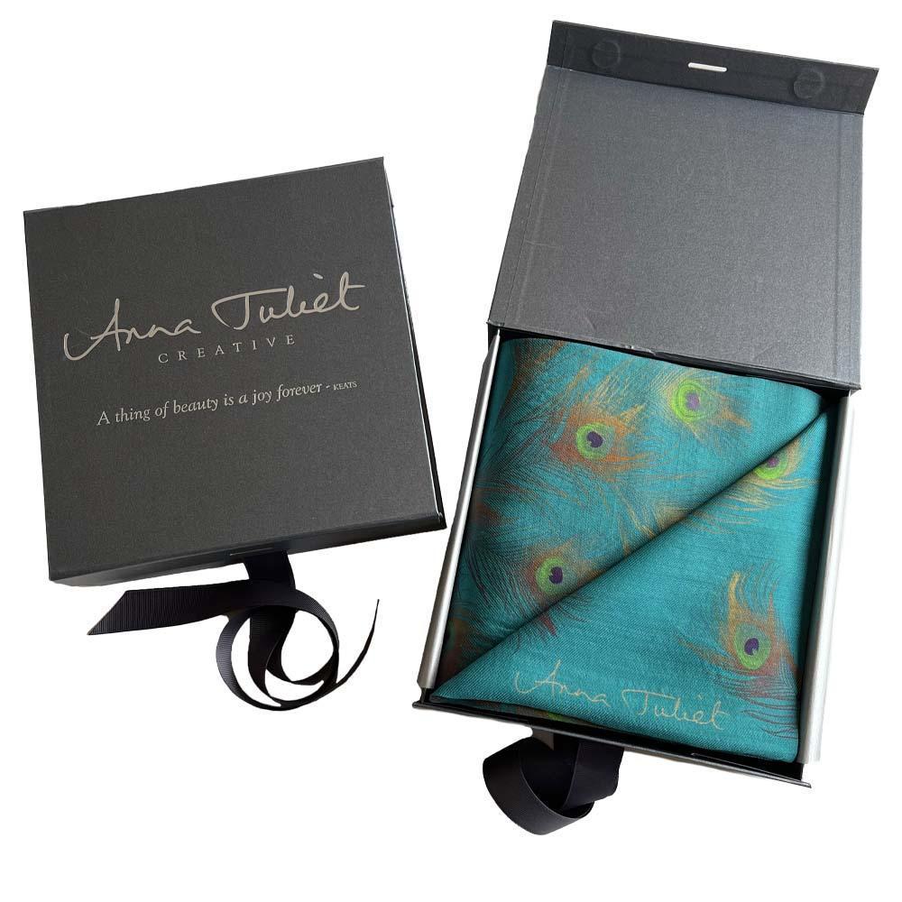 The gift box on the left is closed so that you can see the branding on the pewter-coloured square box. The box on the right is open, with silver tissue paper wrapping open to show the folded wool-silk scarf as it would appear when you open the gift.