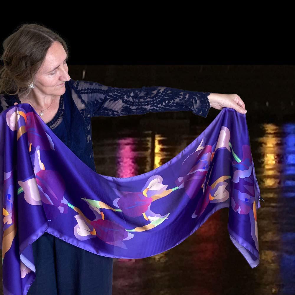 The artist is holding a mauve and blue long silk scarf. In the background coloured lights reflect on water.