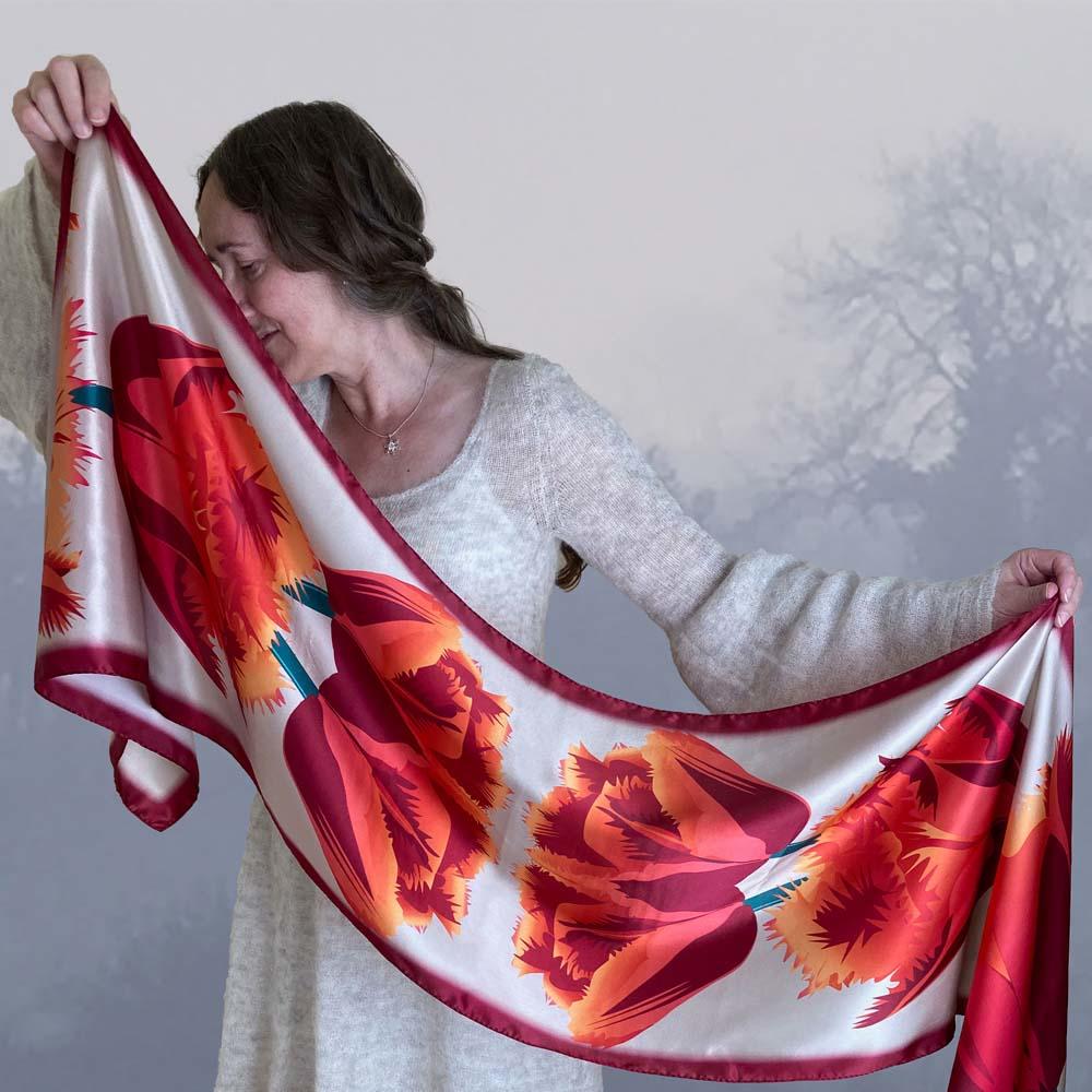 The artist is holding up a beige floral pure silk scarf. Behind her is a misty Autumn view with trees.