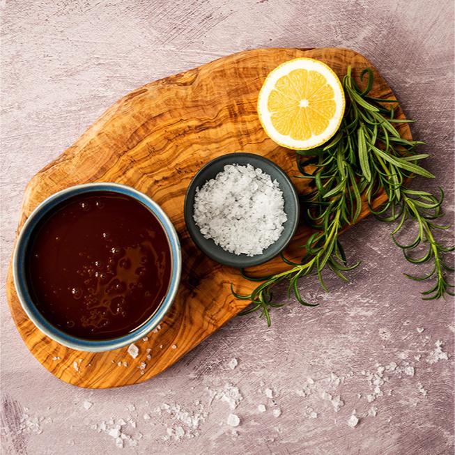 A bowl of Bay's Kitchen Beef stock on a wooden board with salt, lemon, and rosemary