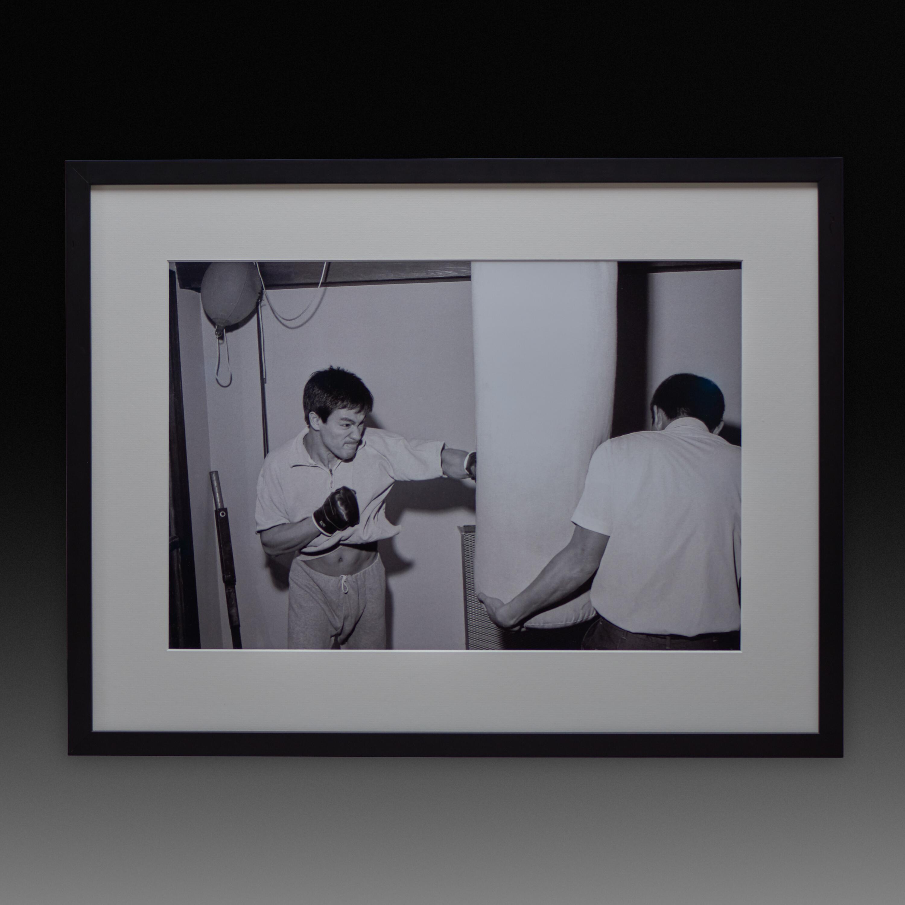 Framed exhibition print of Bruce Lee practicing heavy bag punching with Ted Wong; black and white archival training photo.