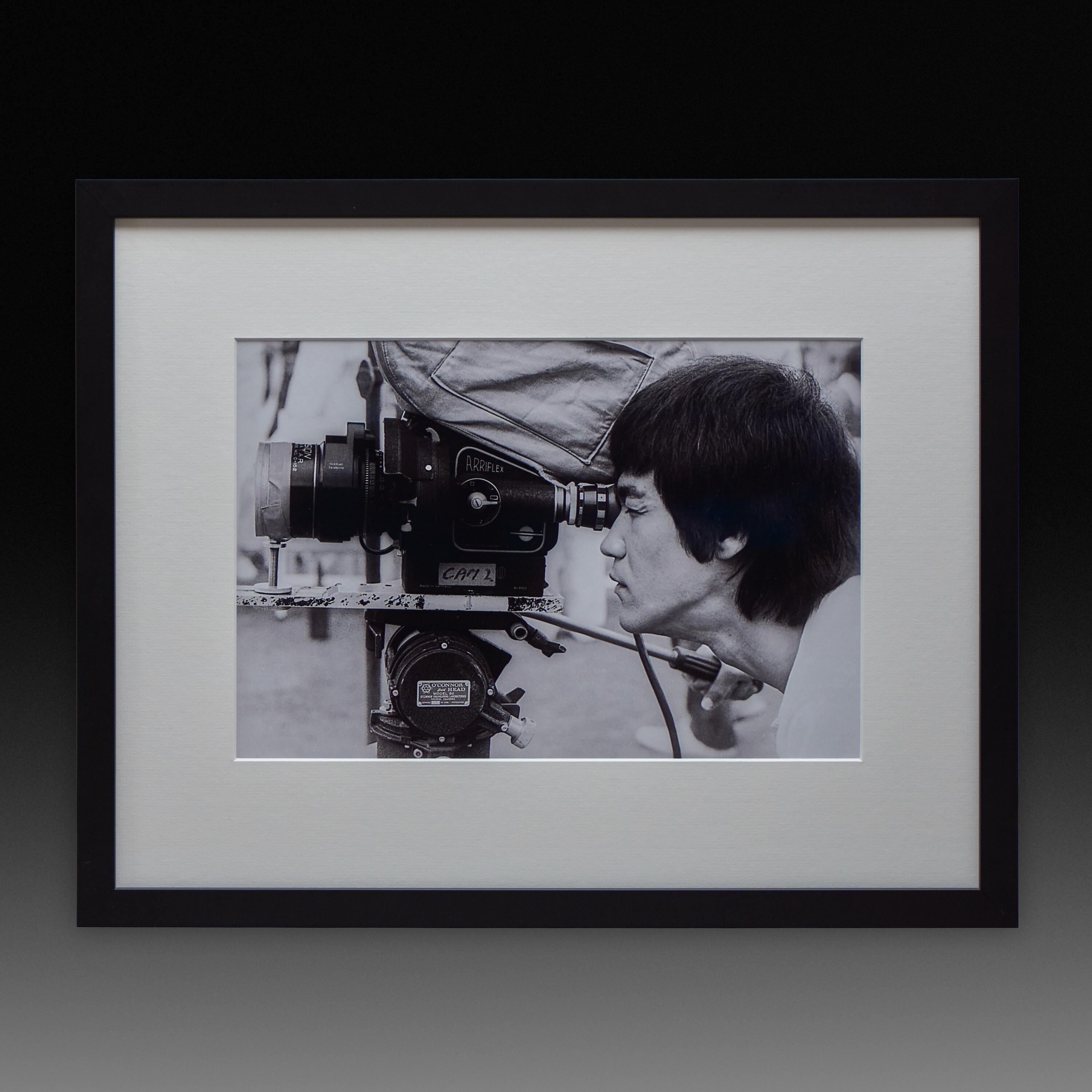 Framed exhibition print of Bruce Lee looking through an Arri flex camera on the Enter the Dragon film set, 1973.