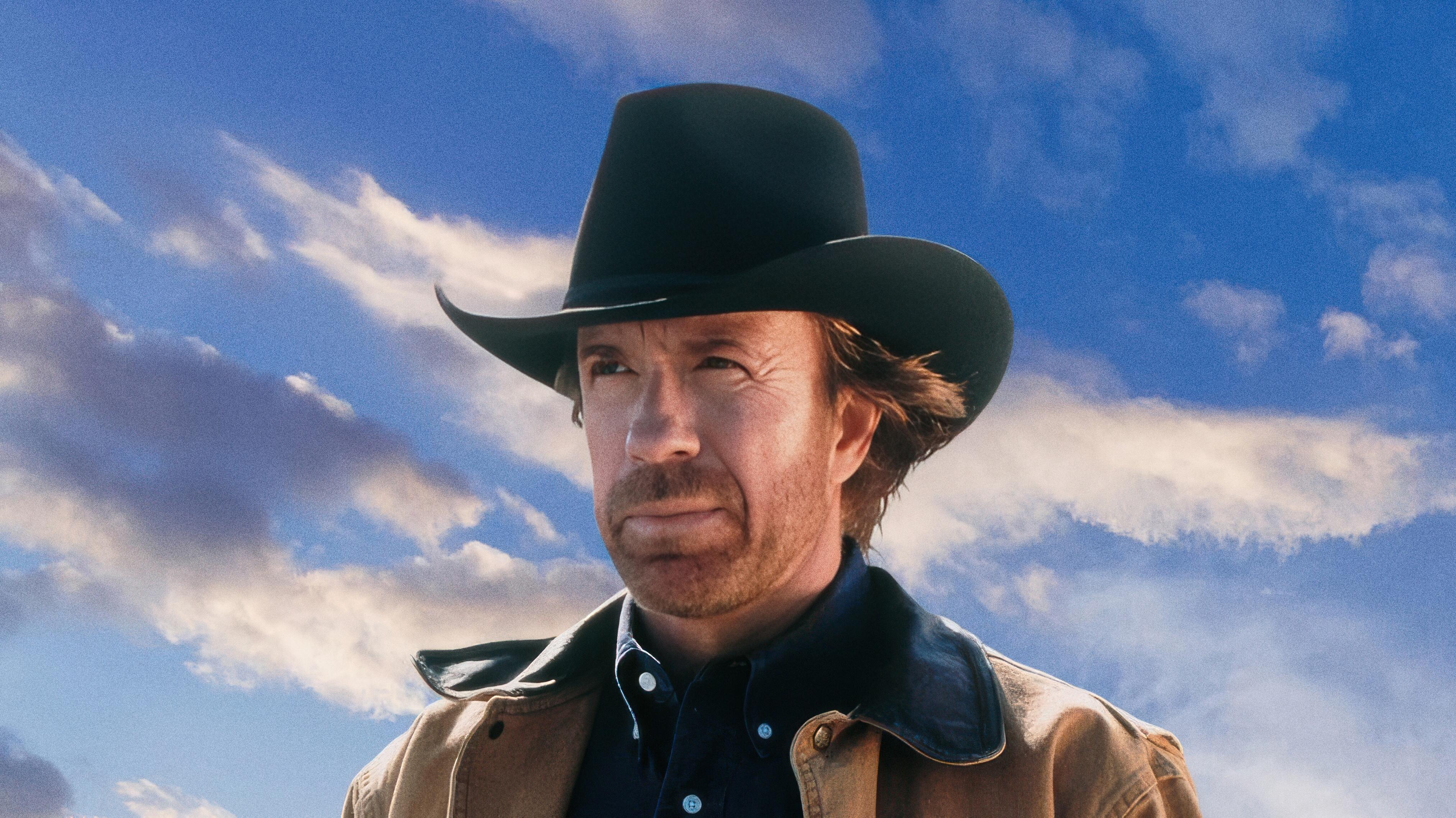 Action star Chuck Norris in his iconic role from Walker, Texas Ranger, wearing a signature black Stetson-style cowboy hat, a dark blue shirt, and a rugged tan coat, looking off-camera with a serious expression under a vast sky.
