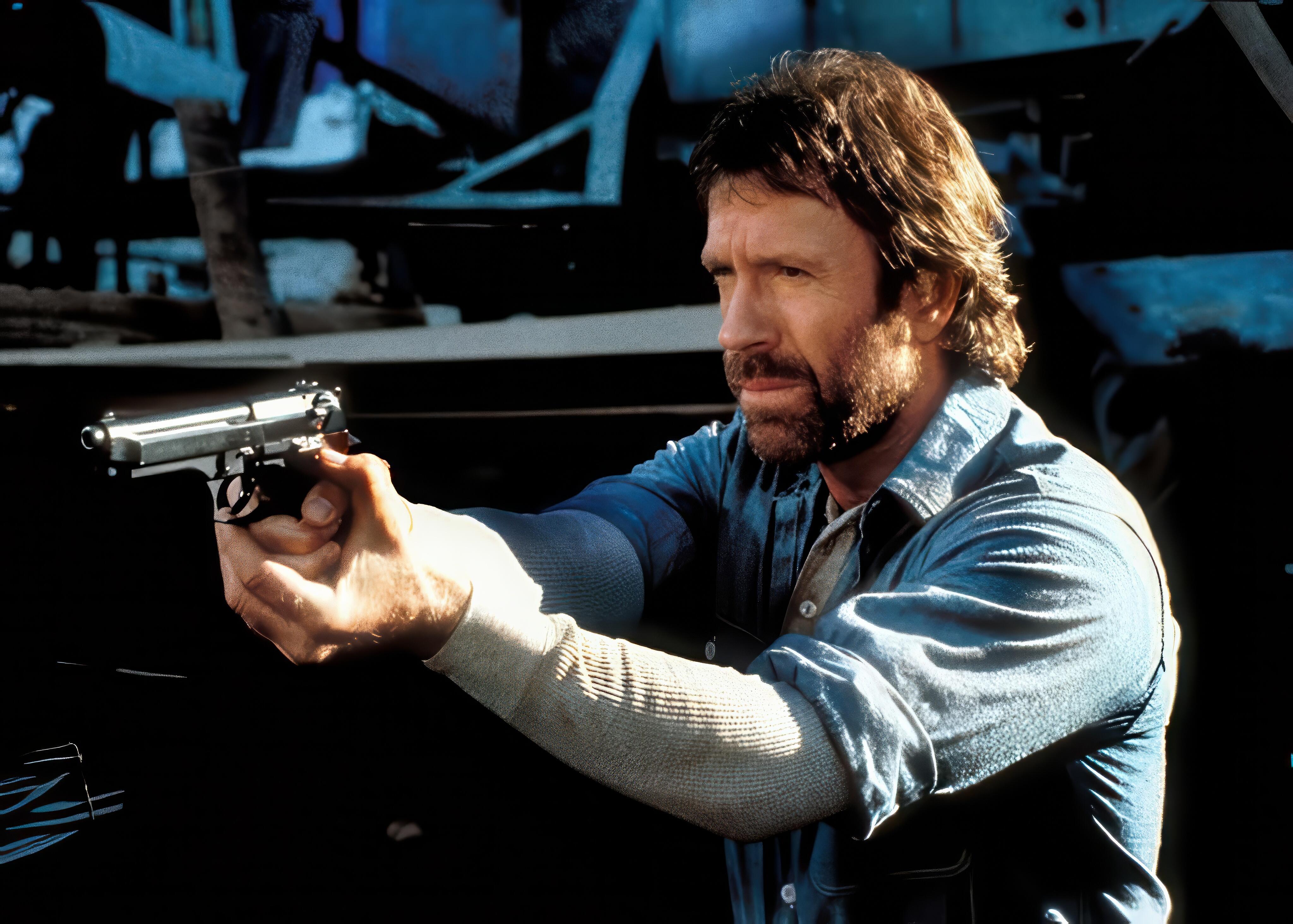 Chuck Norris with his signature beard and blue denim shirt aiming a firearm in the movie ‘Hero and the Terror’ (1988).