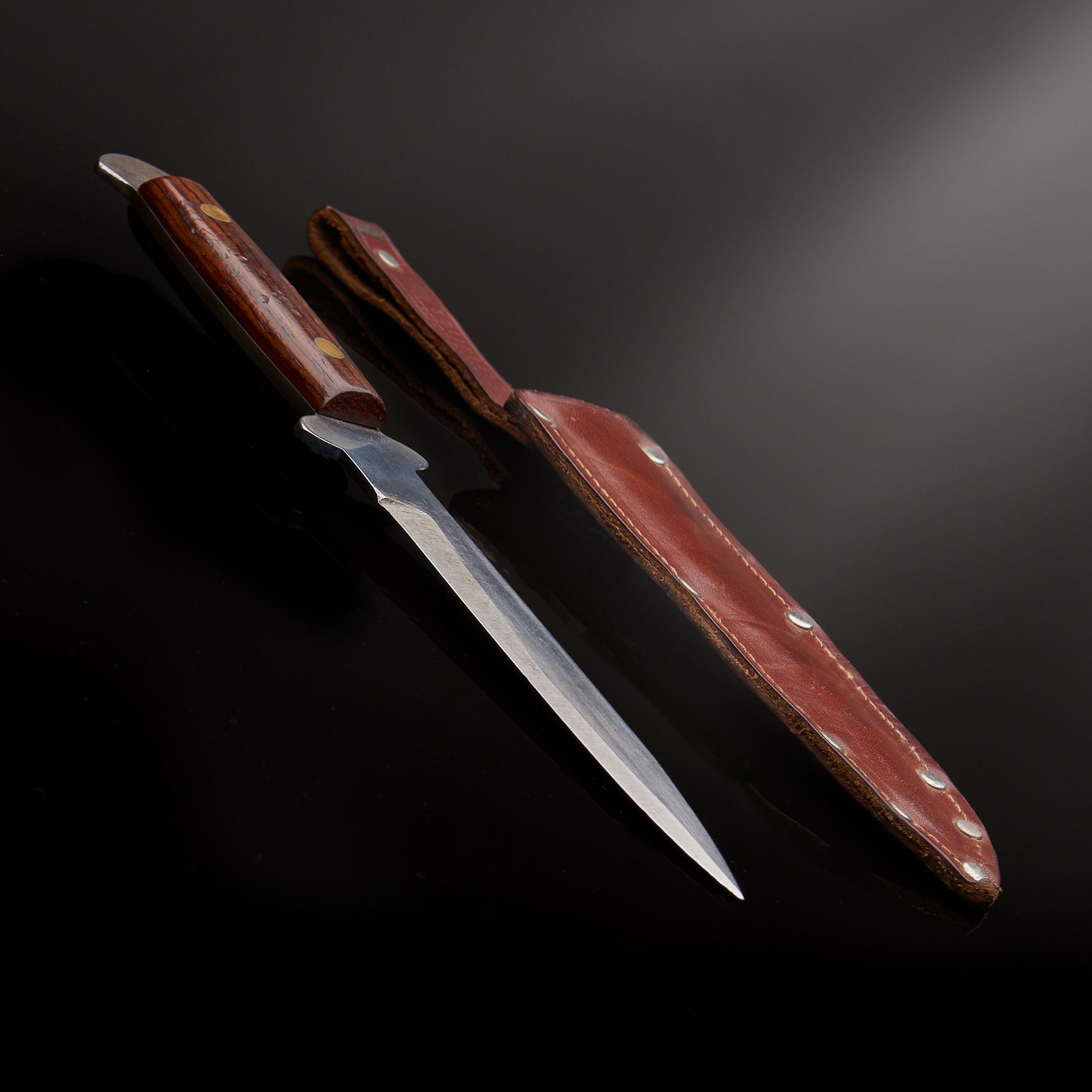 James Bond throwing knife prop from On Her Majesty’s Secret Service, displayed with brown leather sheath on a black surface.