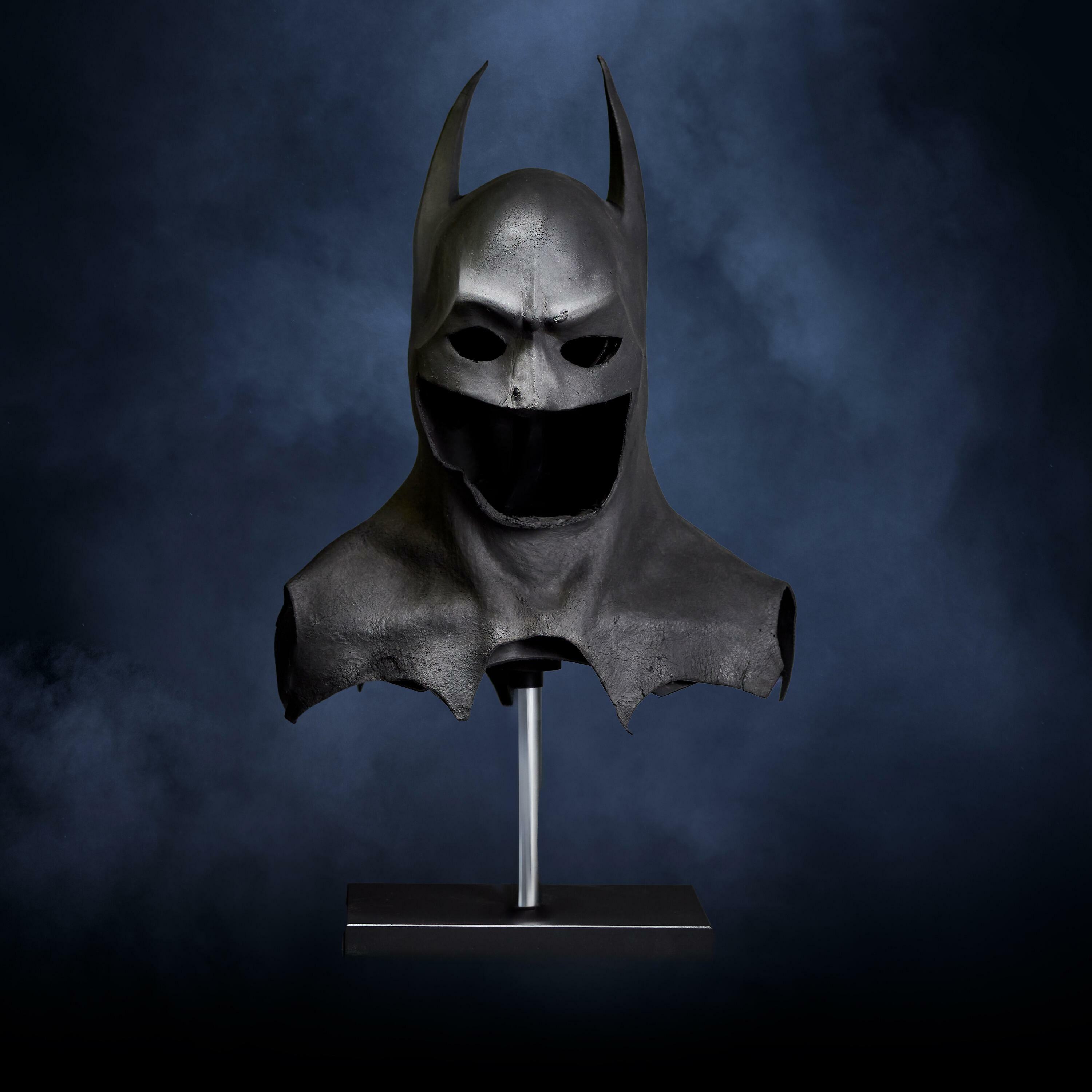 Production-made Batman cowl worn by Michael Keaton, displayed on a stand with a dark blue backdrop