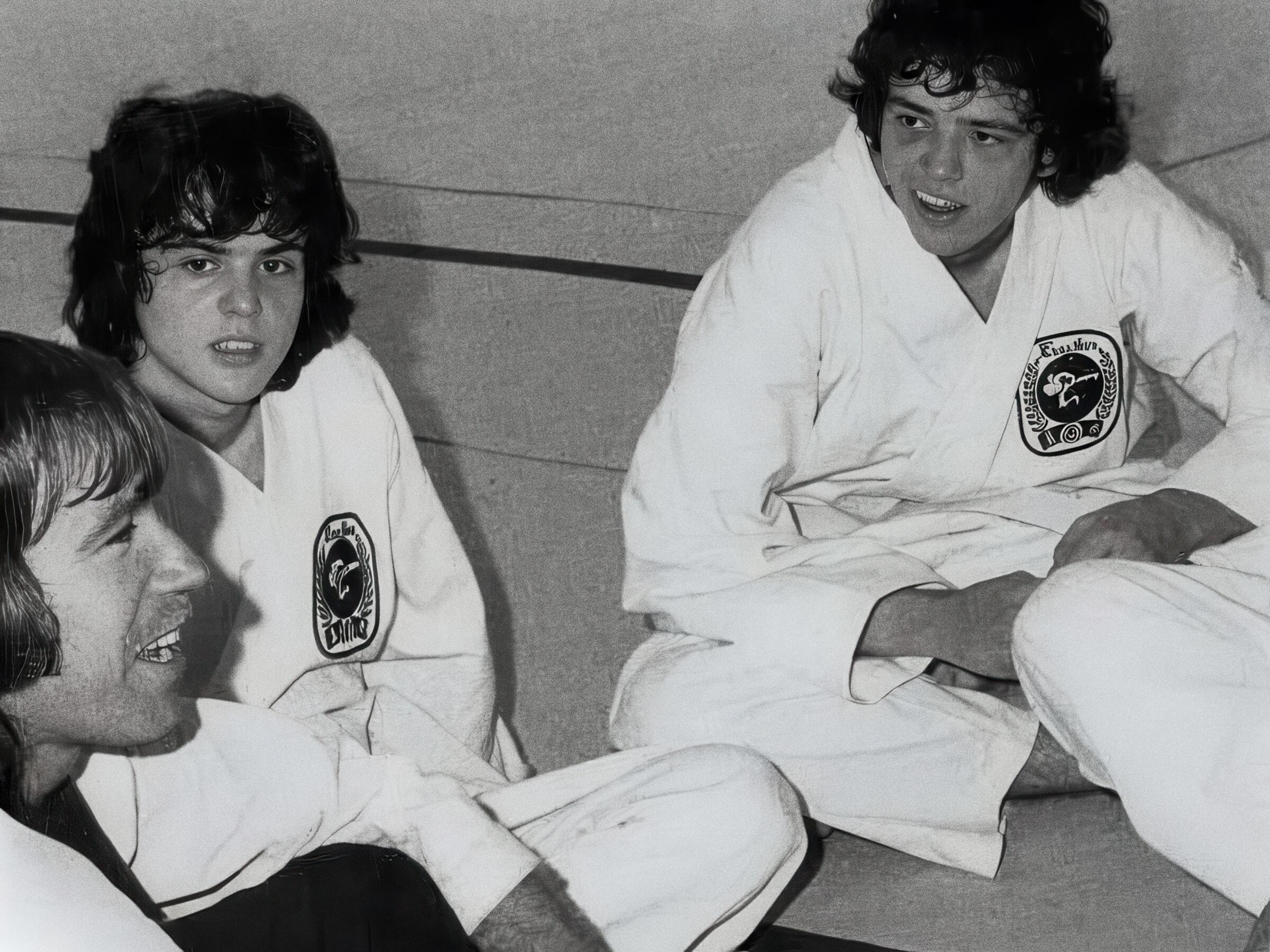 A candid vintage black and white photograph of Chuck Norris laughing during a break in a martial arts training session with members of the Osmond family, all wearing traditional white Tang Soo Do uniforms with circular patches.