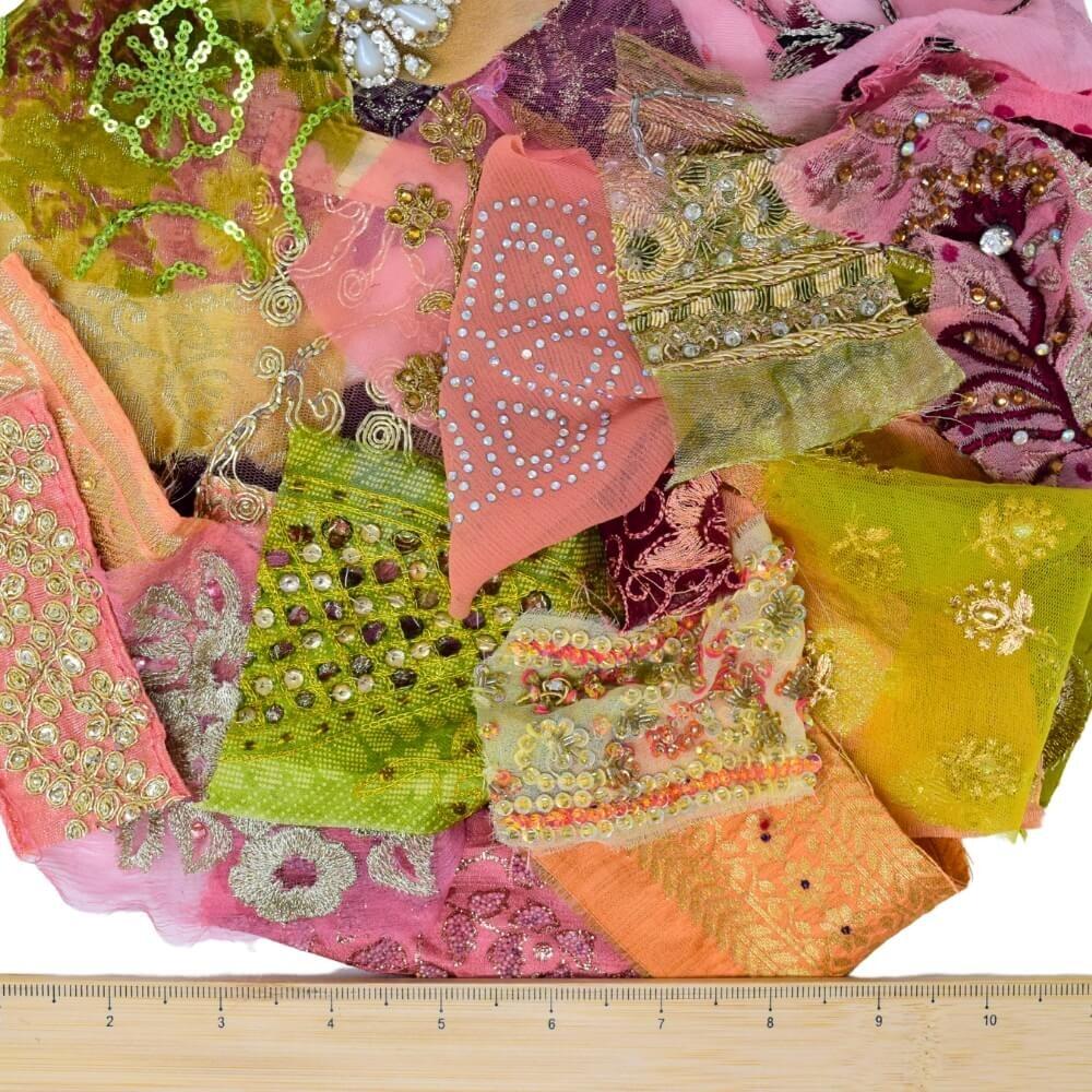 A selection of very small plum, pear and pink coloured, embellished sari fabric scraps with a wooden ruler placed along the bottom edge
