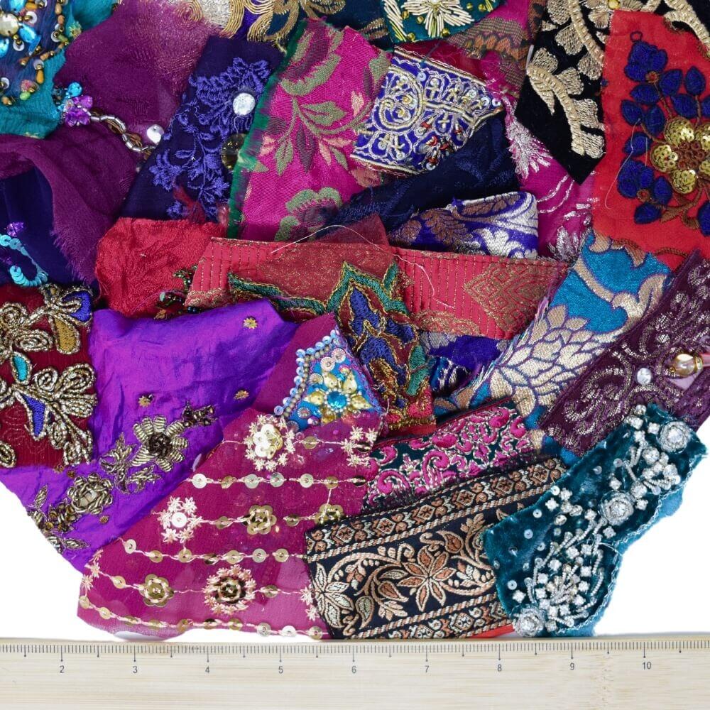 A selection of very small, opulent coloured embellished sari fabric scraps with a wooden ruler placed along the bottom edge