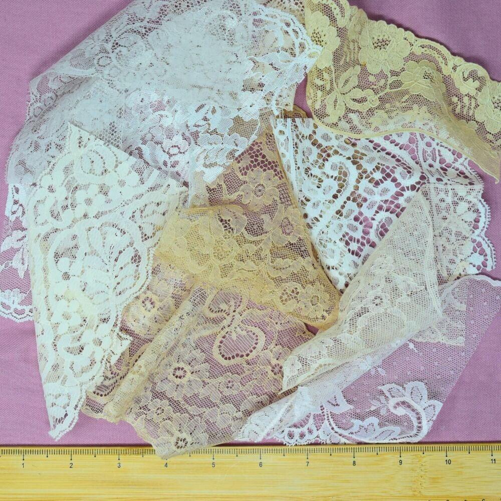 An assortment of lace trims and fabrics arranged in a loose pile on a lilac background with a wooden ruler placed along the bottom edge