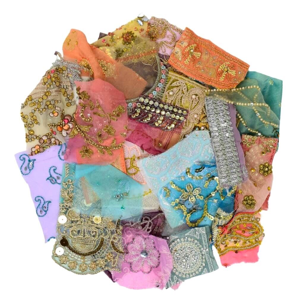 Flat lay of very small pieces of pastel coloured embellished sari fabric scraps arranged in a rough circular shape on a white background