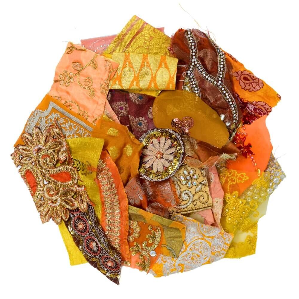 Flat lay of very small pieces of warm coloured embellished sari fabric scraps arranged in a rough circular shape on a white background