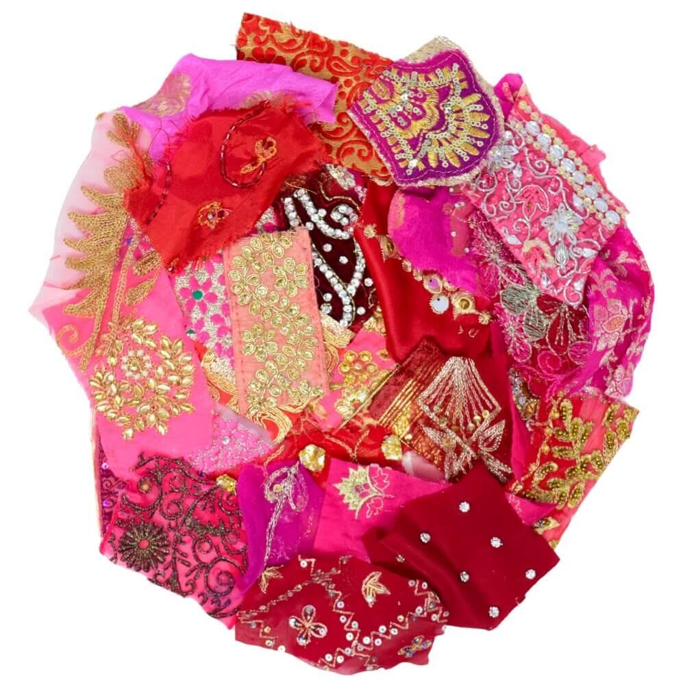 Flat lay of very small pieces of pink and red embellished sari fabric scraps arranged in a rough circular shape on a white background
