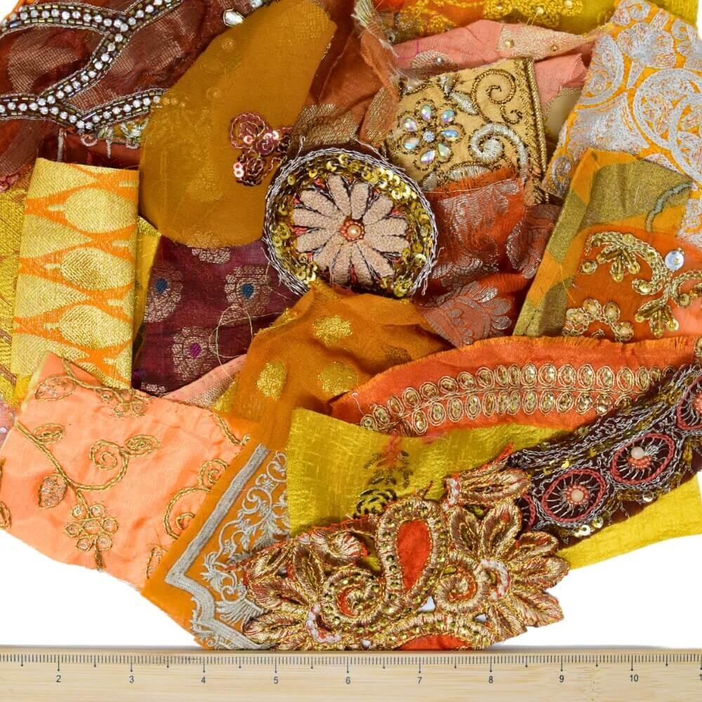 A selection of very small warm coloured, embellished sari fabric scraps with a wooden ruler placed along the bottom edge
