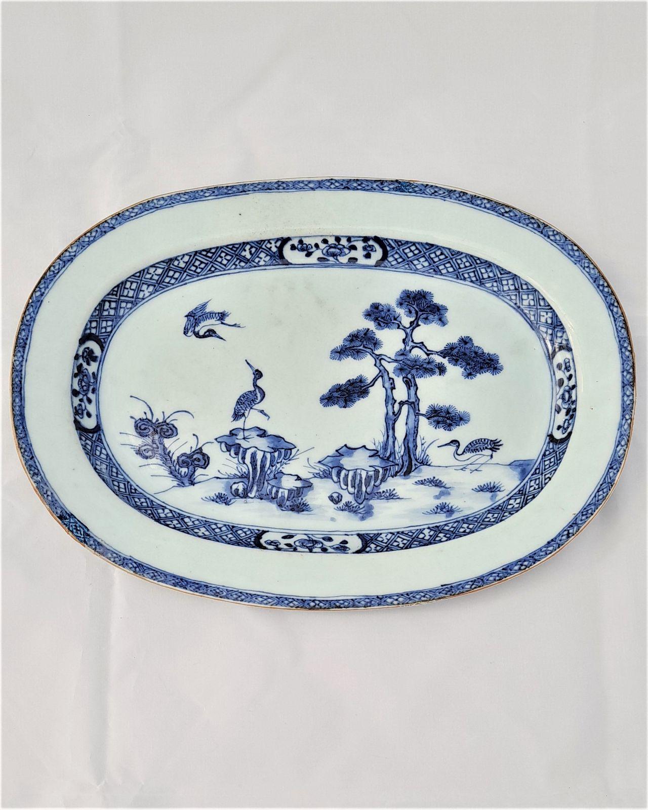 A small oval shaped 12 inch long antique Chinese export porcelain platter, meat plate, serving dish or small charger with hand painted decoration in under-glaze cobalt blue and white with a brown Batavian rim. The beautifully and confidently hand painted design in the centre of the plate has three cranes, one in flight, one amongst rocks, two pine trees and Lingzhi fungus.