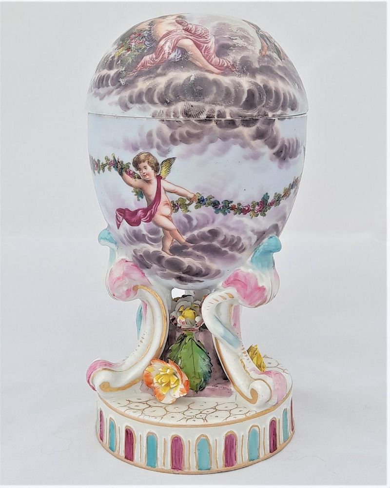 An antique Victorian period mid to late 19th century, ornate hand painted egg shaped porcelain trinket box on rococo scroll tripod legged and floral decorated base.  The egg is hand painted with a scen of Diana and cupid with cherubim. The base is marked underneath with an underglaze blue fish and T mark used by the factory of Carl Thieme of Pottschappel, Frietal, near Dresden, Saxony in Germany circa 1864-1888