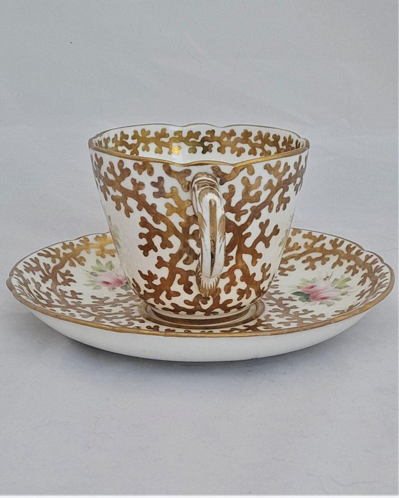 An antique mid Victorian porcelain tea cup and saucer with a scalloped rim and reinforced handle hand painted with roses and gilded extensively with a sea weed pattern made by Sir James Duke & Nephews circa 1860 in the factory previously run by Samuel Alcock and company.