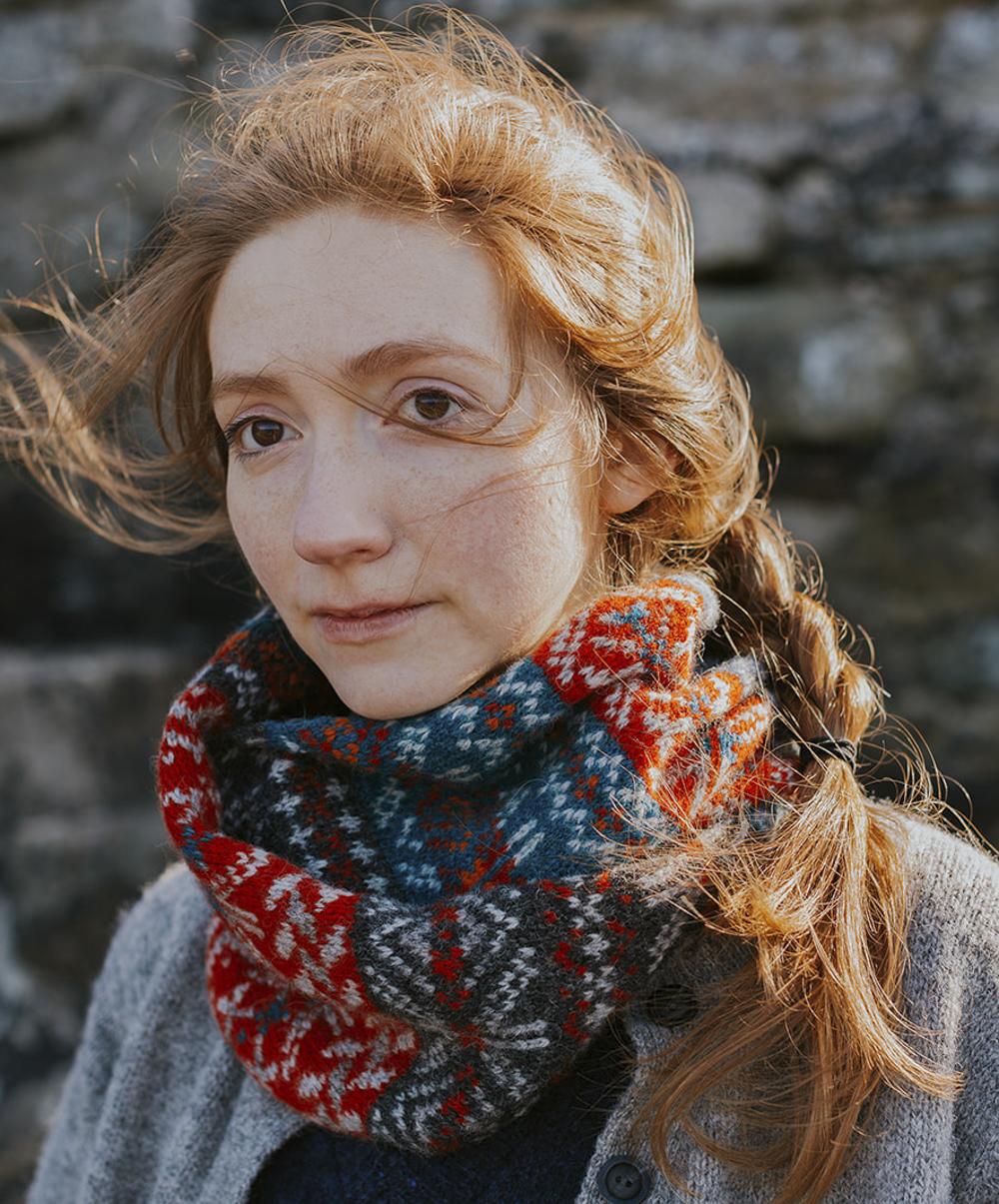 A woman with red hair looks into the middle distance, she is backlit by the sun and wears a richly-patterned Fair Isle cowl which is folded up snugly around her neck