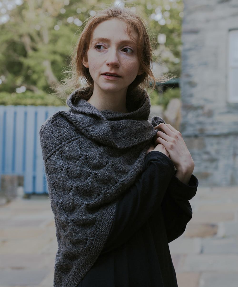 A woman with red hair is all wrapped up in wool, wearing a graphite-coloured deep grey lacy shawl, worked in natural sheepy shades of grey