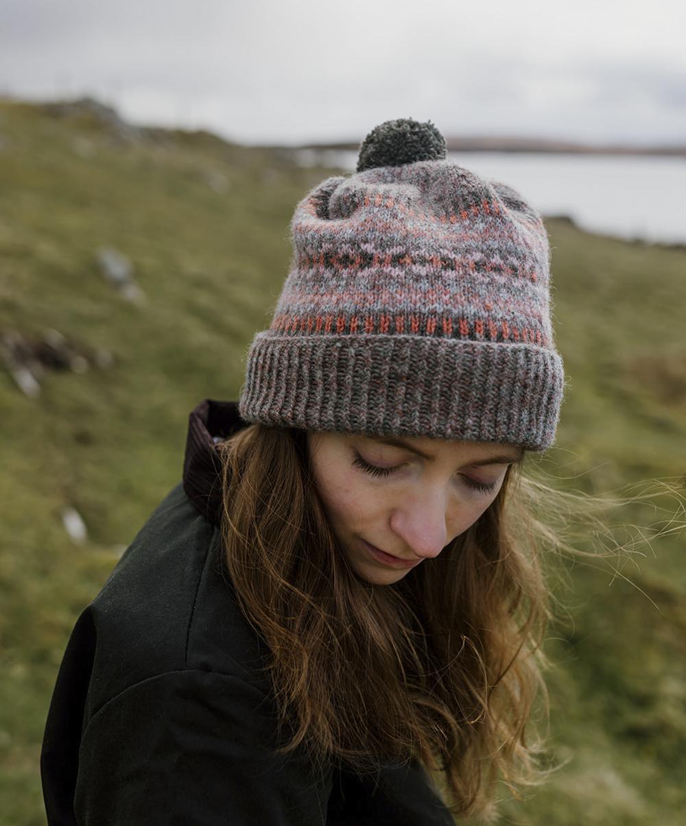 A woman with red hair gazes downwards with a cliff behind her; on her head she wears a richly-patterned hat in shades of grey and pink