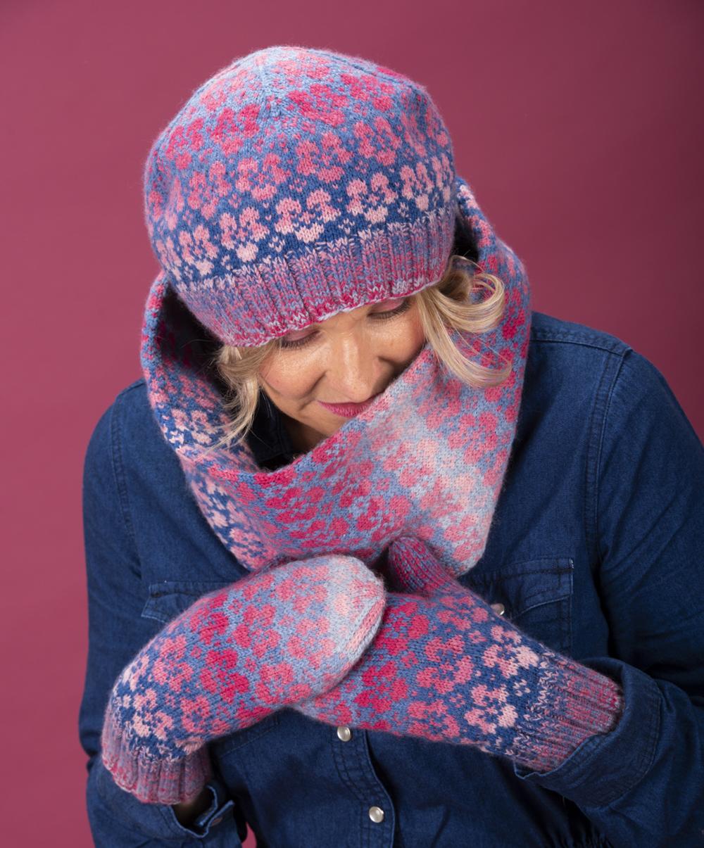 Heidi wears the full Flombre set - matching hat, cowl and mittens each worked in gradients of pink flowers against blue grounds; the background in the image is pink and Heidi wears a deep blue denim dress with the hand knitted Flombre accessories