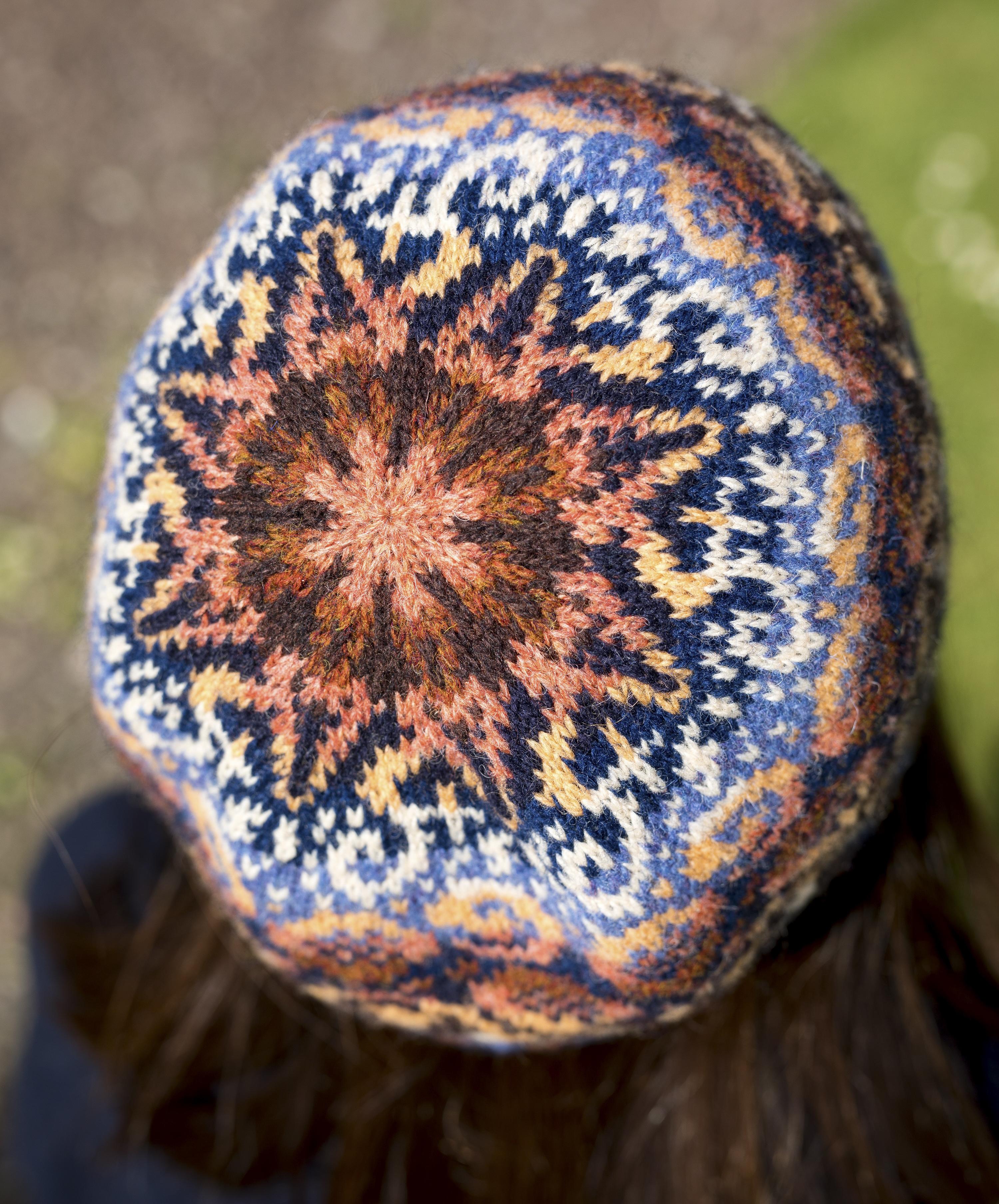 The crown of a stranded colourwork hat, in which the stitches decrease evenly in spokes towards the centre of the hat. Worked in a palette of corals, golden shades and deep warm blues and browns.