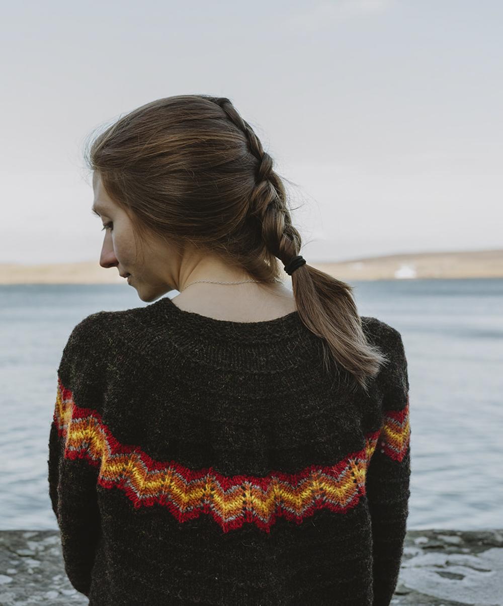 A woman with red hair stares out to sea while wearing a black gansey (sweater) with a striking fire-inspired yoke detail - Up Helly Aa gansey by Susan Molloy