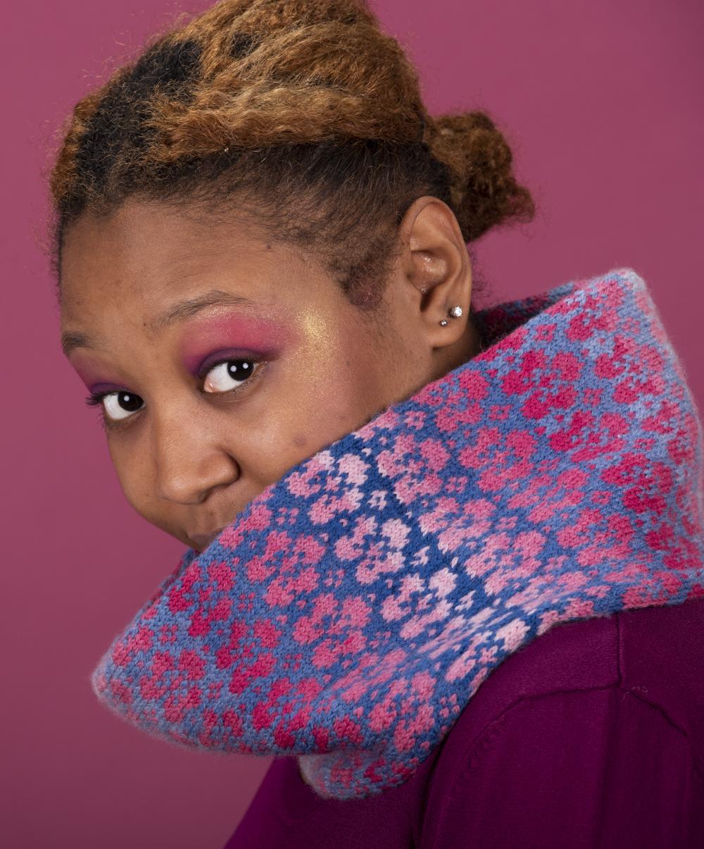Taia peeps out of the Flombre cowl; it is worked with a blue gradient in background and pink gradient flowers as the pattern and Taia wears an ombre top in deep to mid pink; the background of the photo is also pink