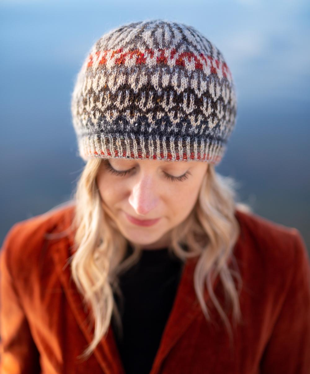 Jane Hunter Models Featherheid - a stranded colourwork hat design based on duck plumage; the background is blue with lochs and skylines