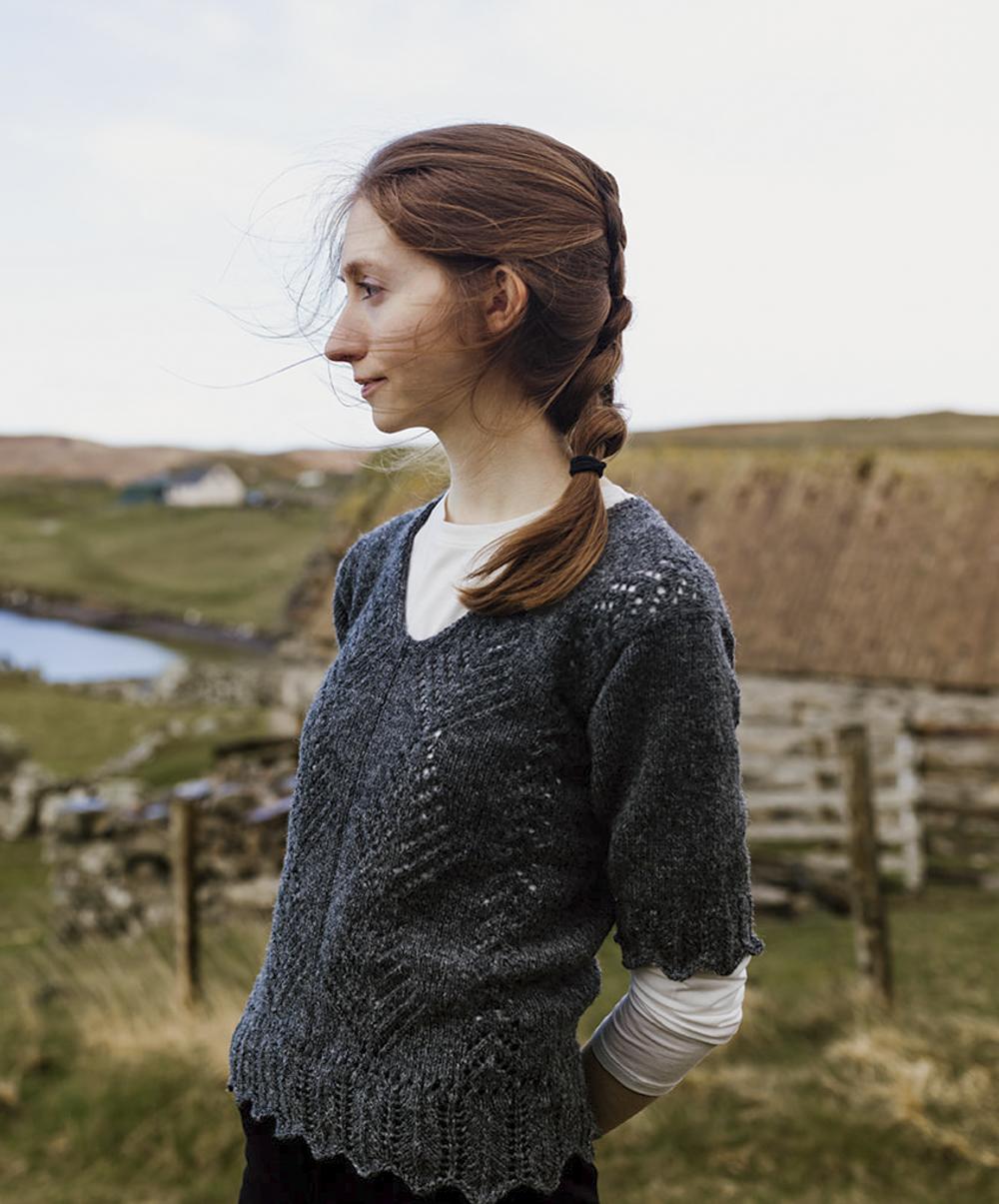 A woman with red hair stares off into the middle distance, behind her is a thatched croft house and she is wearing a lacy sheepy tunic, the colour of graphite