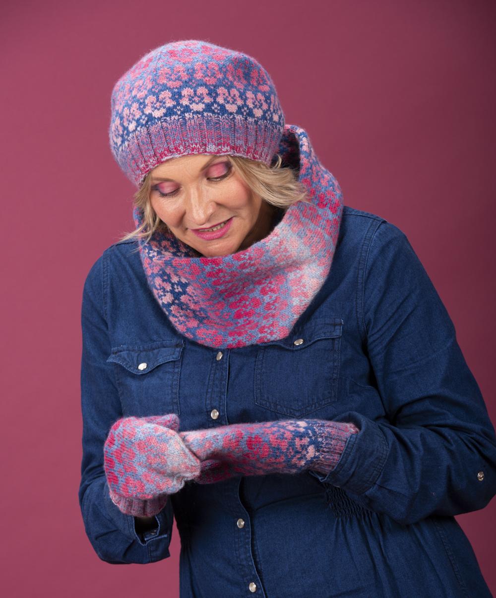 Heidi examines the pattern on one of the Flombre mittens; she wears the matching pink and blue accessories and a dark blue denim dress; the background is pink