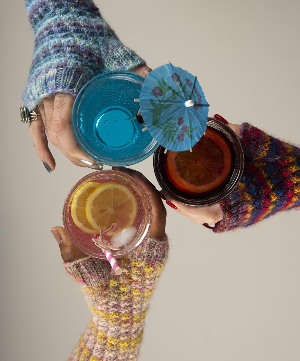 Three hands - each wearing a differently-coloured version of the Cheers! mitts - toast their glasses together; each edition of the mitts is clearly inspired by the cocktail in the glass the wearer holds