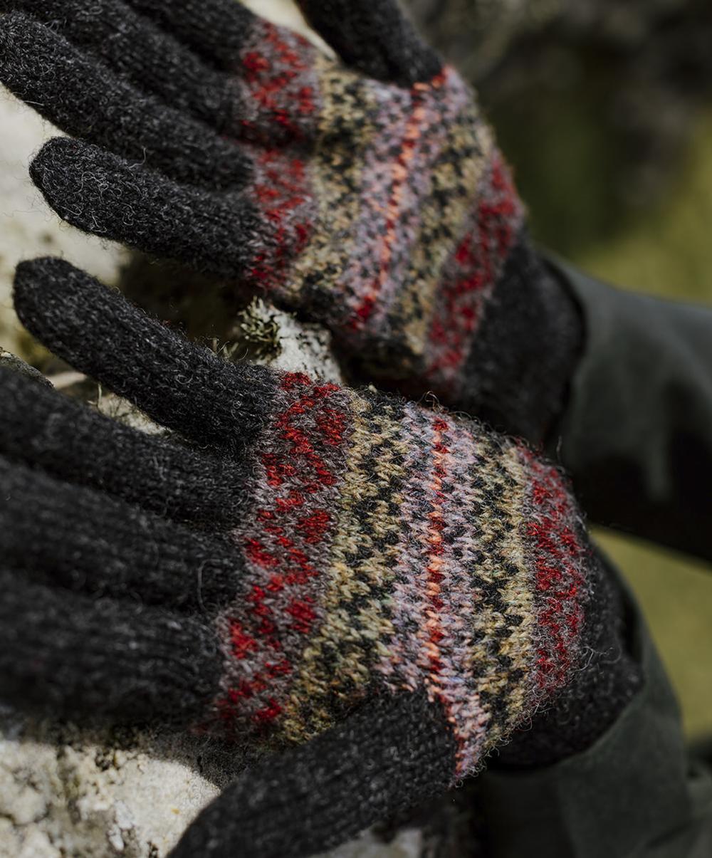 Hands are open on a lichen-covered wall; the hands wear gloves in a deep charcoal shade with richly-patterned hands and palms