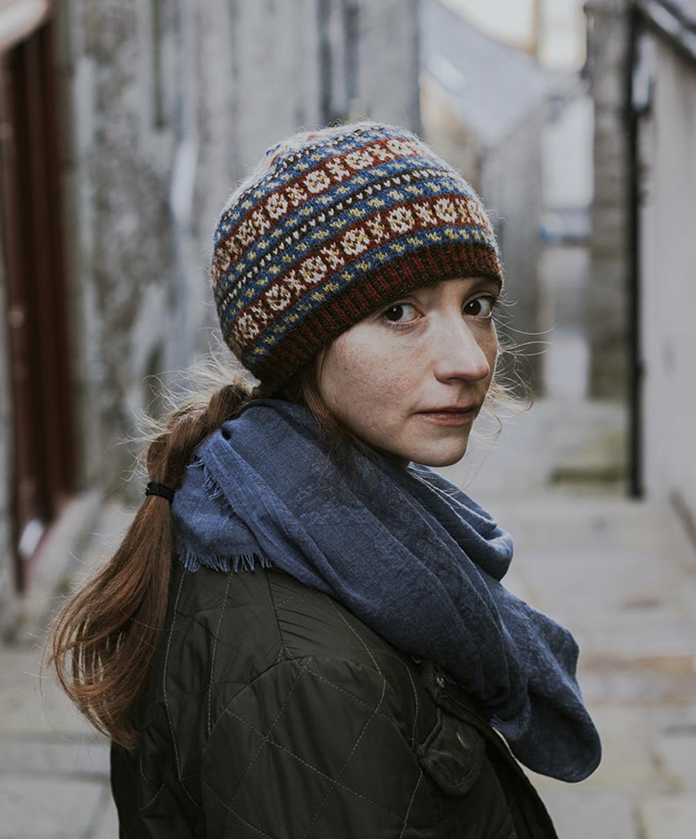 A woman with red hair turns to face the camera while wearing a richly-patterned Fair Isle beanie featuring small (peerie) patterns in the traditional palette of dark reds, indigo blues, golds and browns and greys which would have been used by Shetlanders prior to widespread availability of synthetic dyes