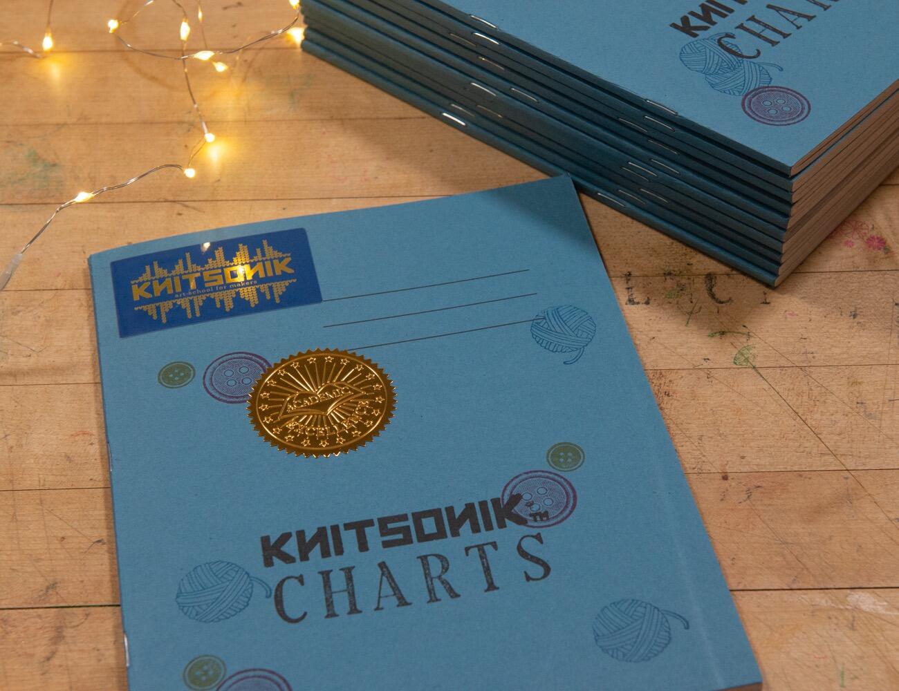 KNITSONIK notebook - a plain grey blue exercise book, customised with sparkly stickers and rubber stamp impressions, lying on a wooden table with fairy lights beside it