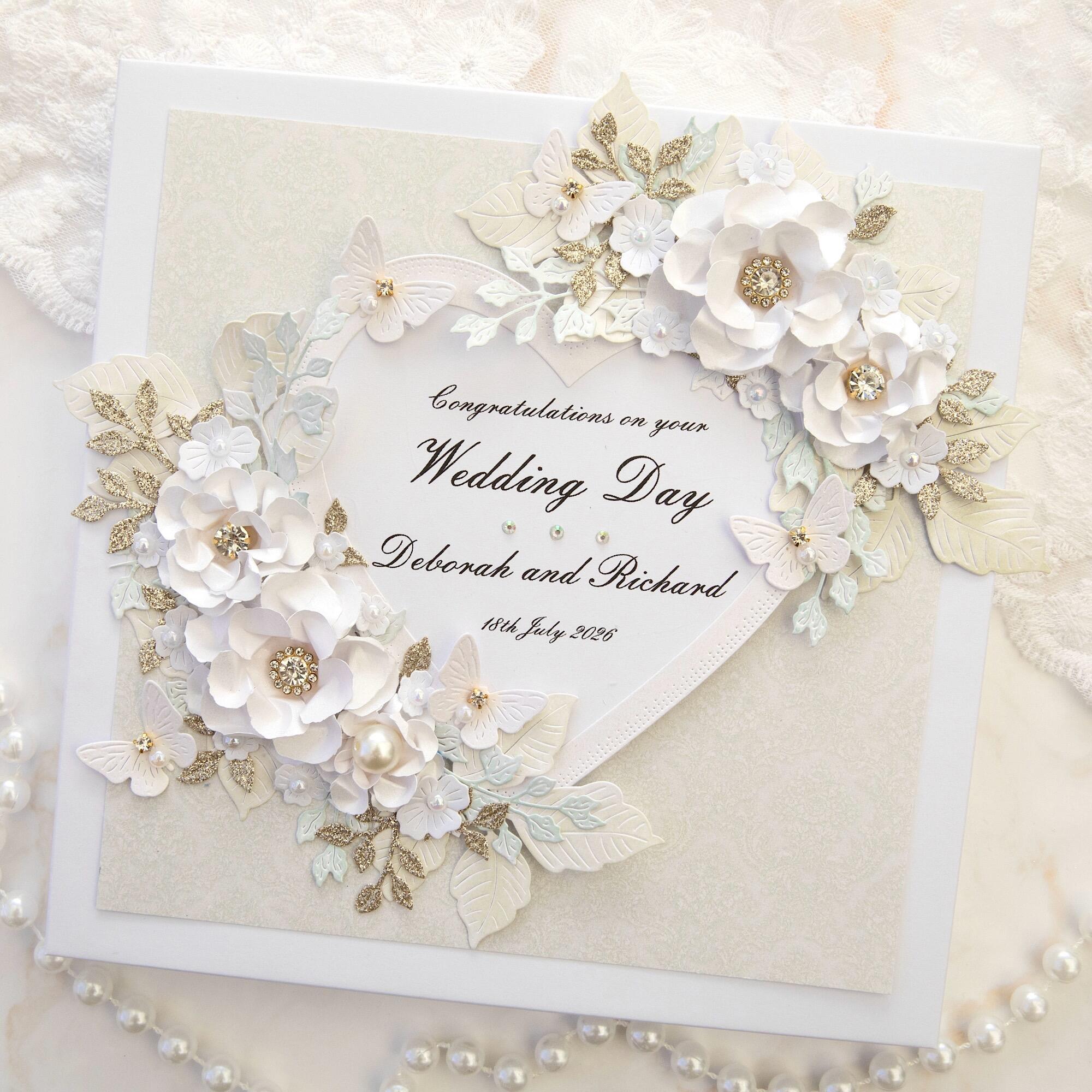 Luxury handmade personalised 8x8 white and gold wedding card with large heart reading Congratulations on your Wedding Day Deborah and Richard 18th July 2026, decorated with 3D white paper flowers with crystal centres, white butterflies, pearls and gold glitter leaves, handmade in Leicestershire.