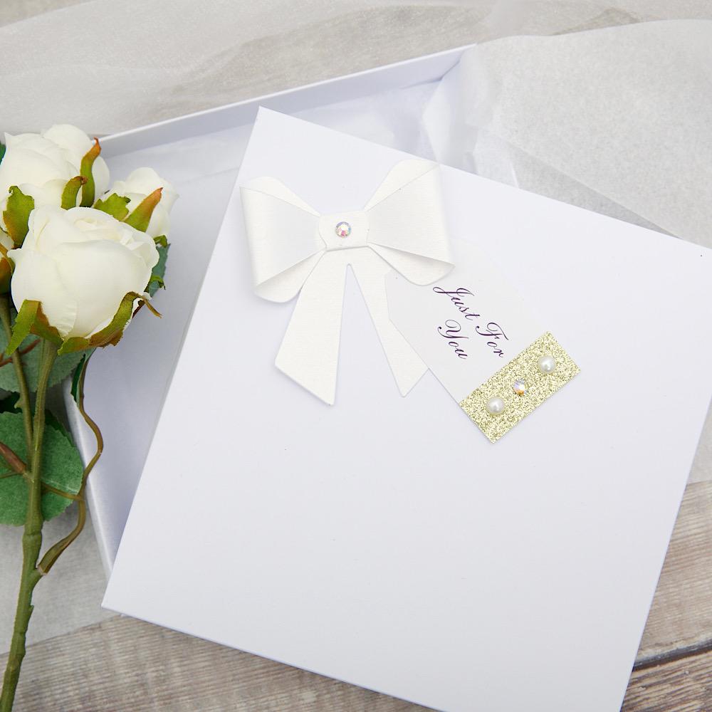 A gift box decorated with white paper bow and tag sits on a wooden background. The lid is partially open, showing tissue paper lining inside the box.